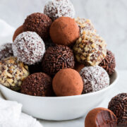 Stack of homemade chocolate truffles covered in cocoa, nuts, sprinkles and coconut piled into a pyramidal shape in a white bowl set over a marble surface.