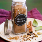 Garam masala spice blend in a glass jar set over a plate with whole spices next to it.