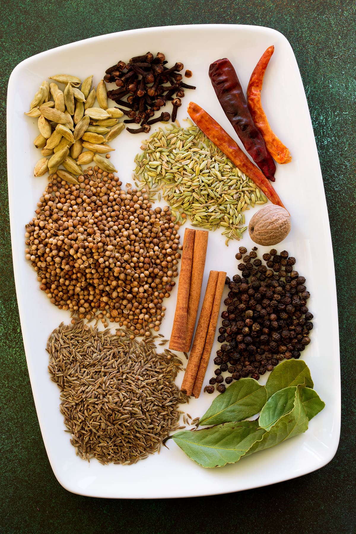 Rectangular white platter with whole spices used to make garam masala. Includes cumin seed, coriander seed, cardamom pods, whole cloves, red peppers, fennel seeds, whole nutmeg, cinnamon sticks, black peppercorn and bay leaves.