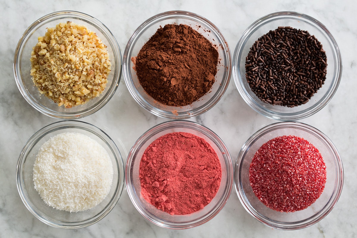 Photo of topping ideas of chocolate truffles. Shows chopped nuts, cocoa powder, sprinkles, shredded coconut, pulverized freeze dried strawberries and colored sugar in individual small glass bowls.