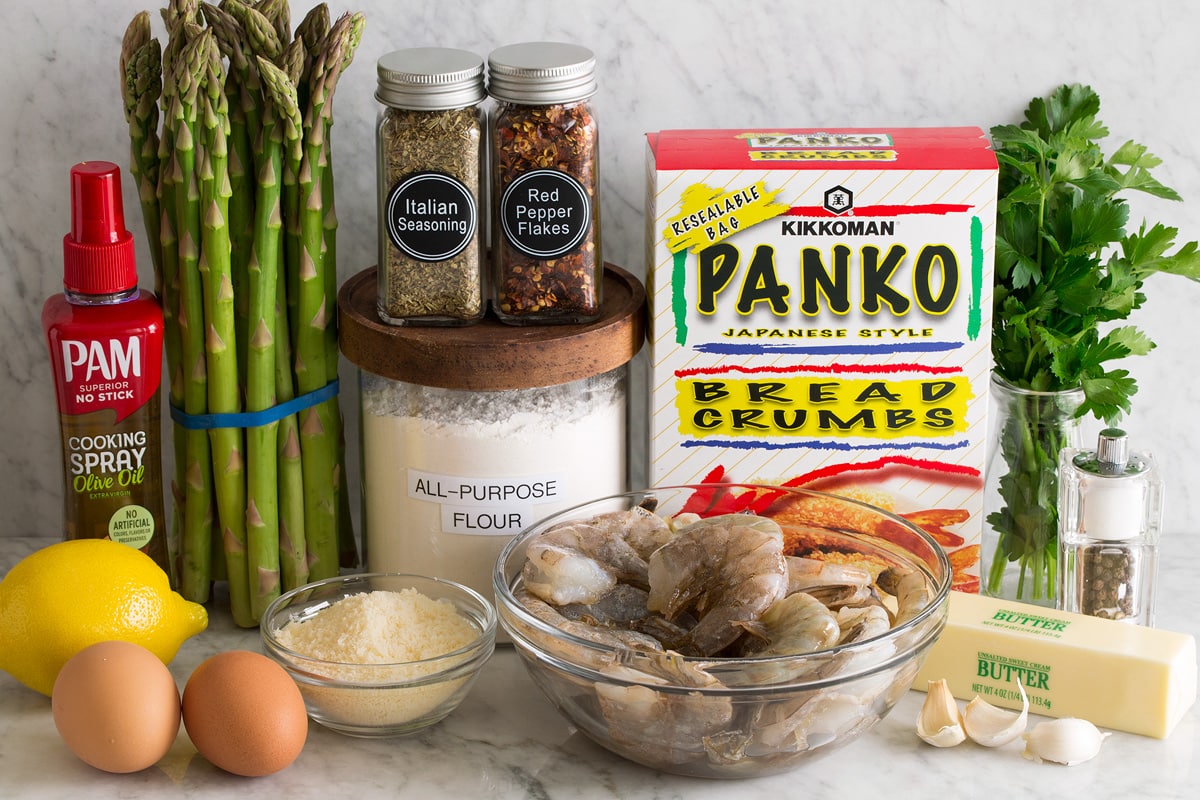 Photo of ingredients for sheet pan shrimp and asparagus. Includes raw shrimp, garlic, butter, salt, pepper, parsley, panko, flour, red pepper flakes, Italian seasoning, parmesan, eggs, lemon, asparagus and olive oil cooking spray.