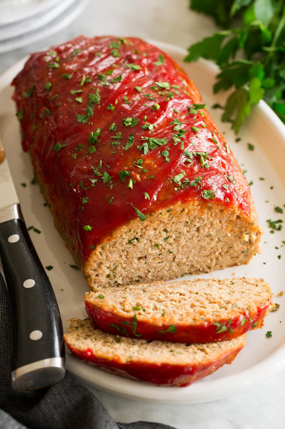 Photo of turkey meatloaf on a white oval serving platter. Parsley is sprinkled over and whole parsley is to the side. A large knife is shown to the side.