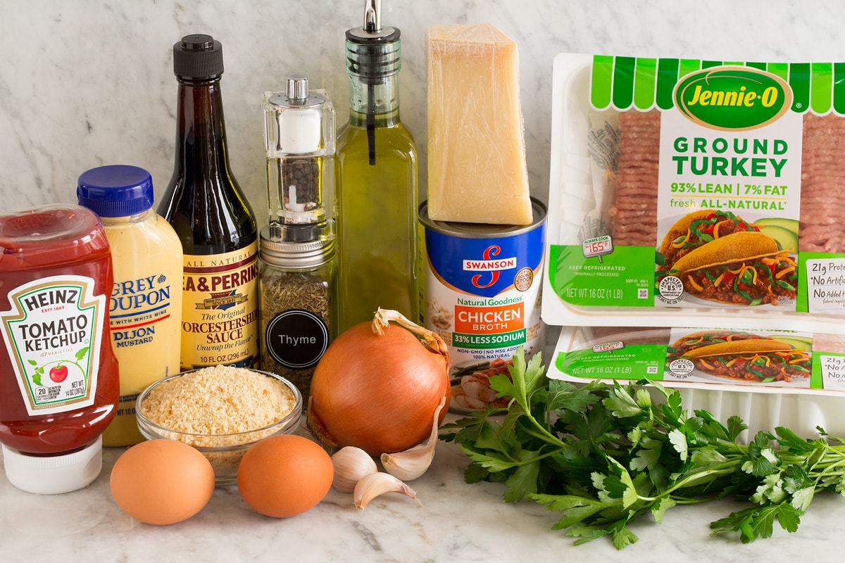 Photo of ingredients used in turkey meatloaf. Includes ground turkey, parsley, parmesan, chicken broth, olive oil, yellow onion, garlic cloves, eggs, bread crumbs, thyme, salt and pepper, Worcestershire, dijon, ketchup. 