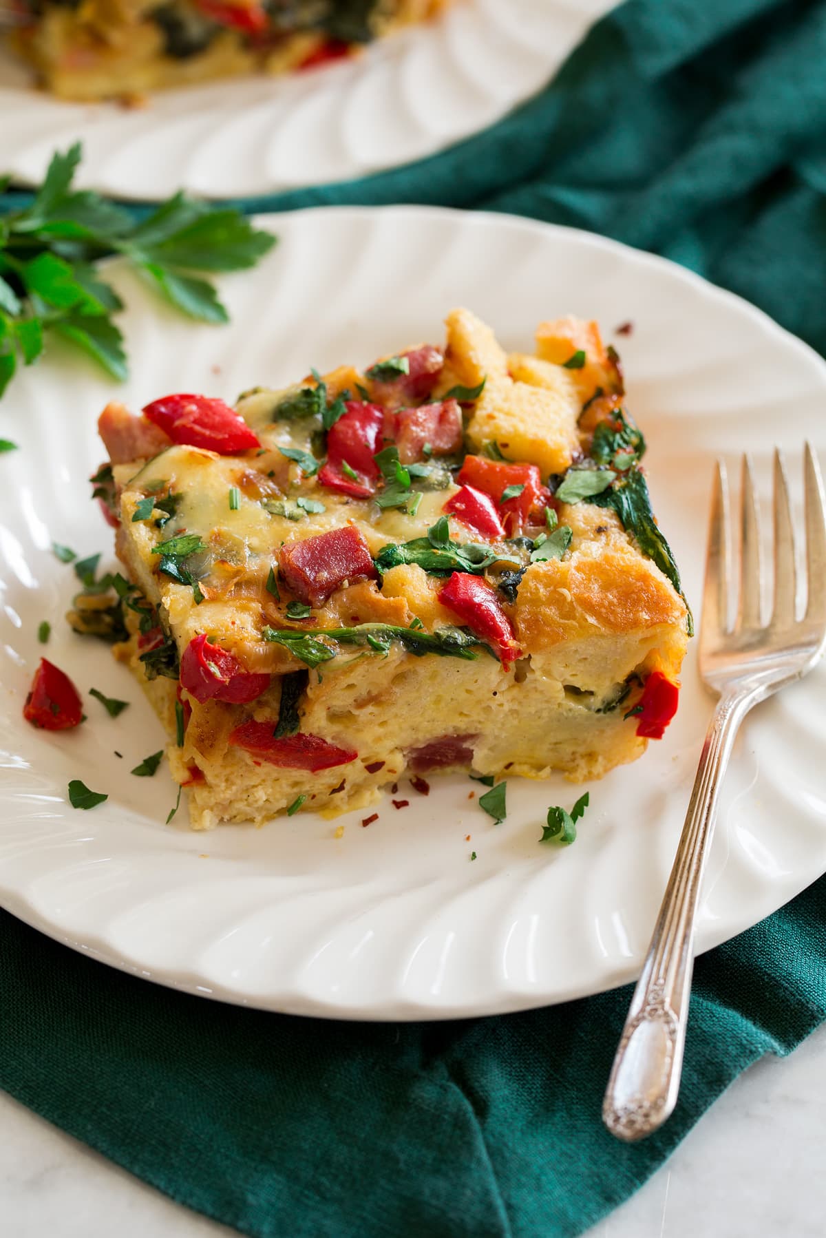 Photo of slice of strata on a single serving plate with a fork to the side. Plate is set over a green cloth.