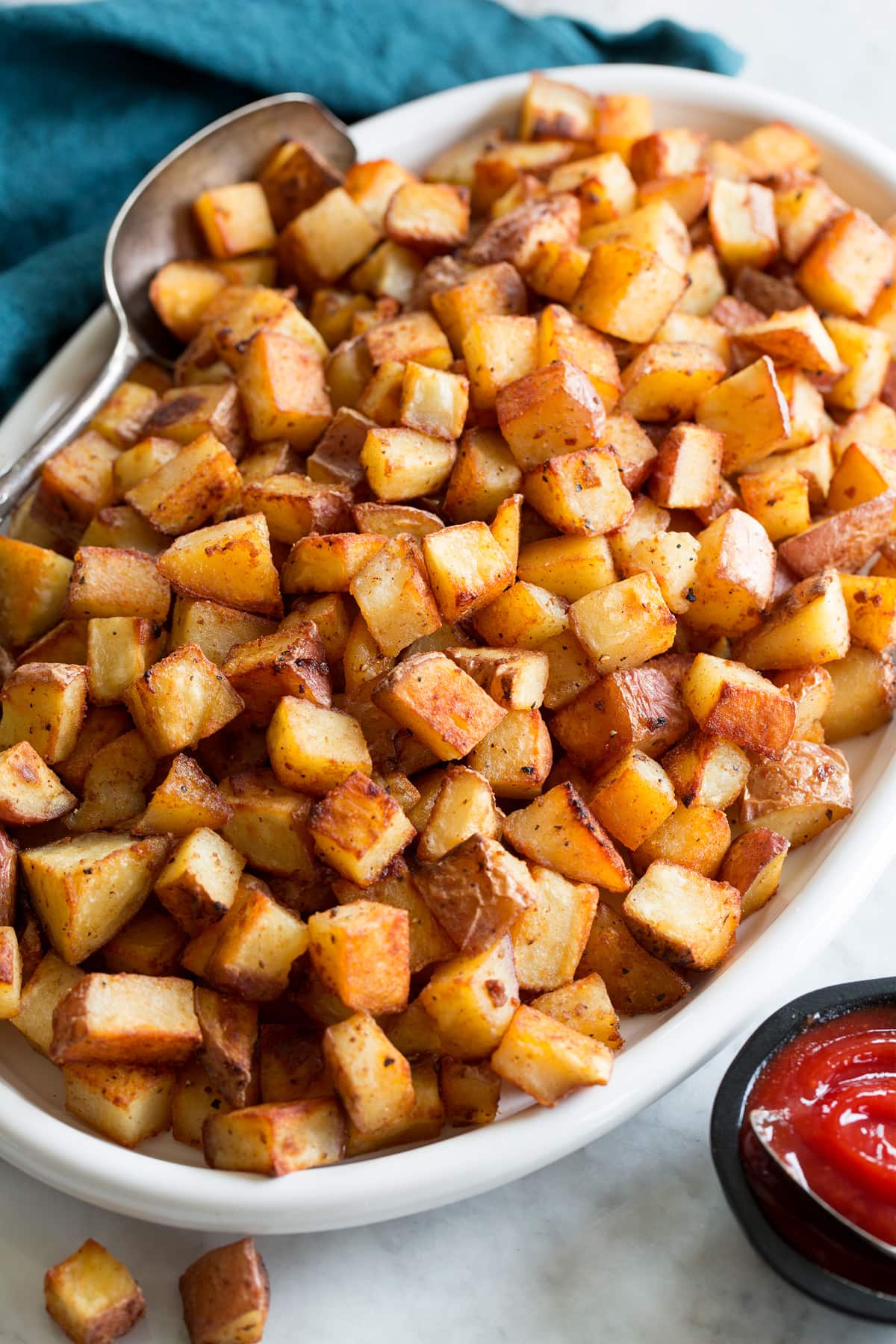 Photo: Breakfast Potatoes on a white oval serving plater with a blue cloth and silver spoon resting to the side. Sriracha ketchup is shown as a serving suggestion.