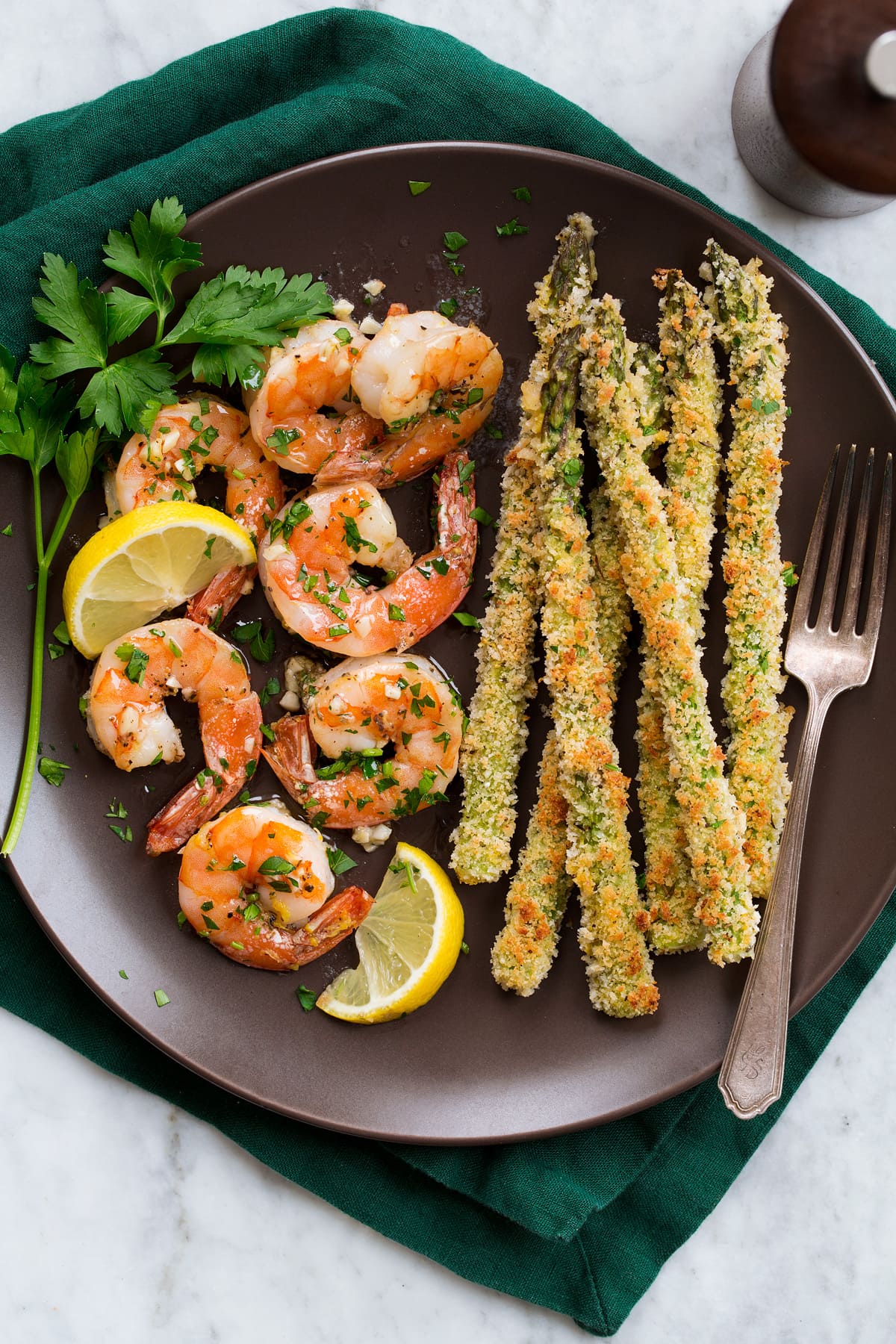 Overhead photo of buttery garlic shrimp and panko asparagus on a dark brown plate set over a dark green cloth napkin.