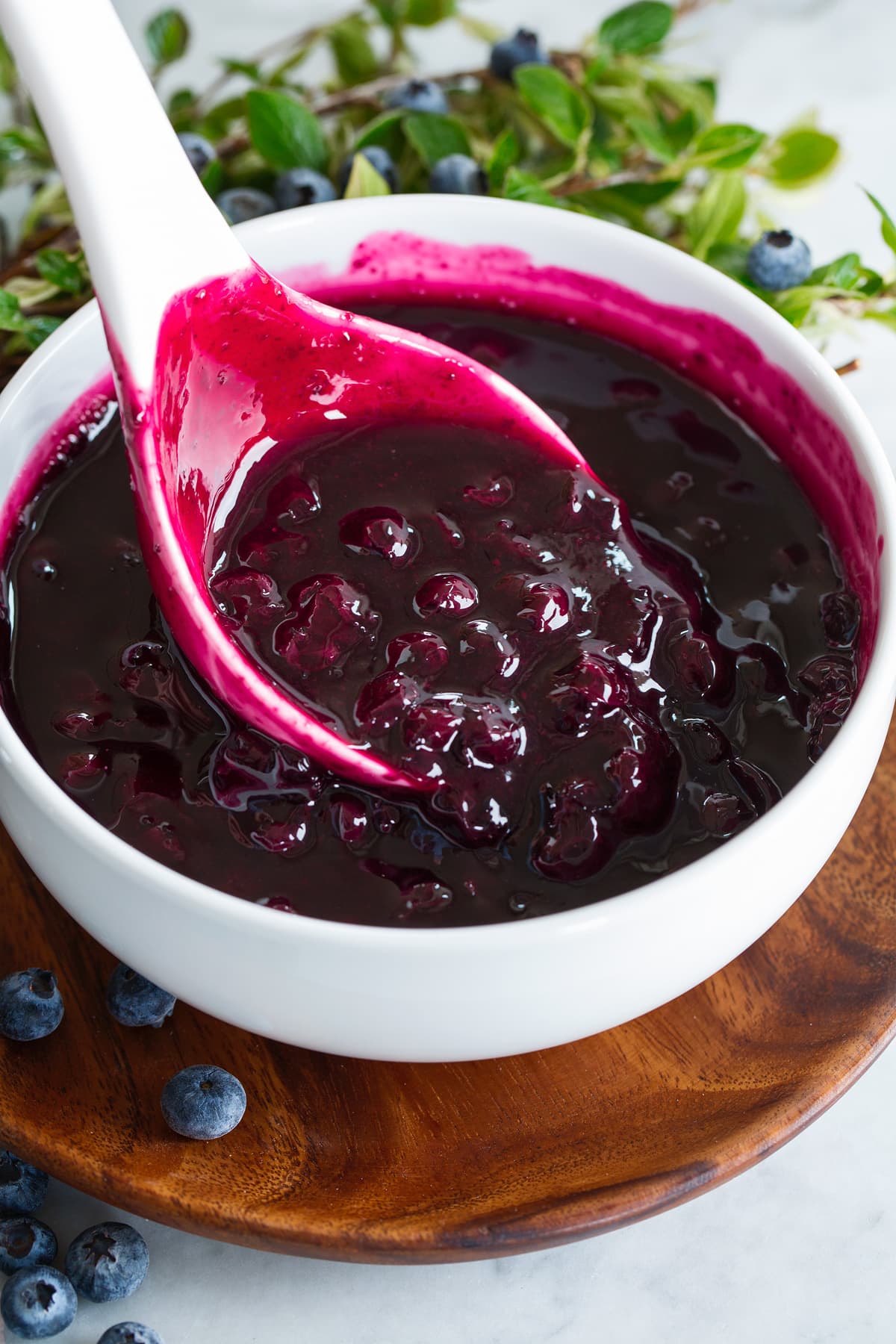 Finished blueberry syrup in a white bowl over a wooden plate. Syrup is being scooped by a large white ladle.