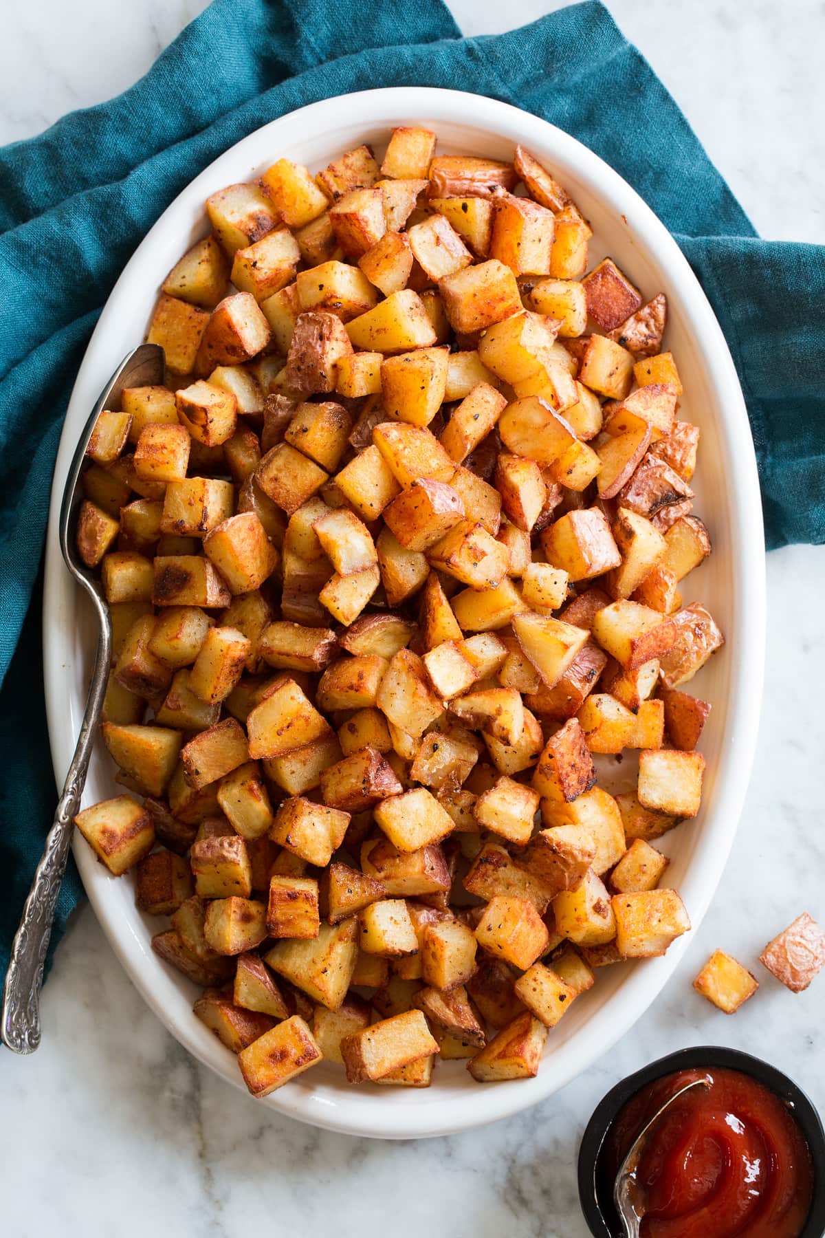 Photo: Diced breakfast potatoes shown from above on a white oval serving platter with a marble surface below and a blue cloth.