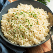 Rice seasoned with garlic and butter shown in a blue bowl over a wooden surface. Rice is topped with pieces of butter and chopped parsley.