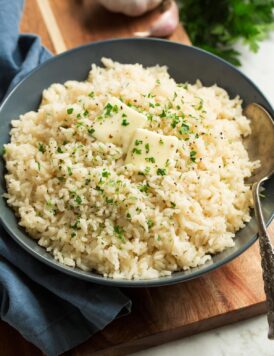 Rice seasoned with garlic and butter shown in a blue bowl over a wooden surface. Rice is topped with pieces of butter and chopped parsley.