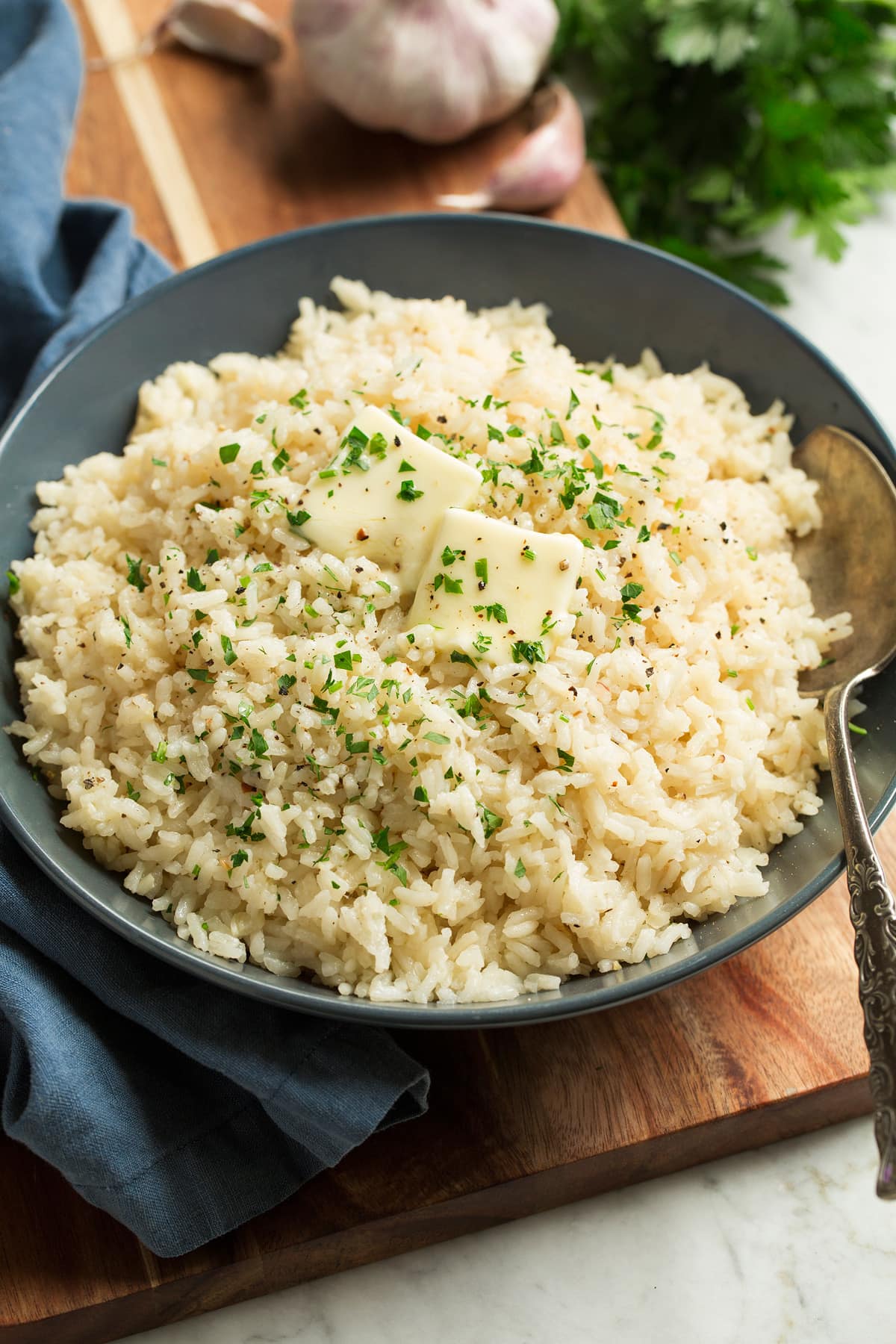 Rice seasoned with garlic and butter shown in a blue bowl over a wooden surface. Rice is topped with pieces of butter and chopped parsley.