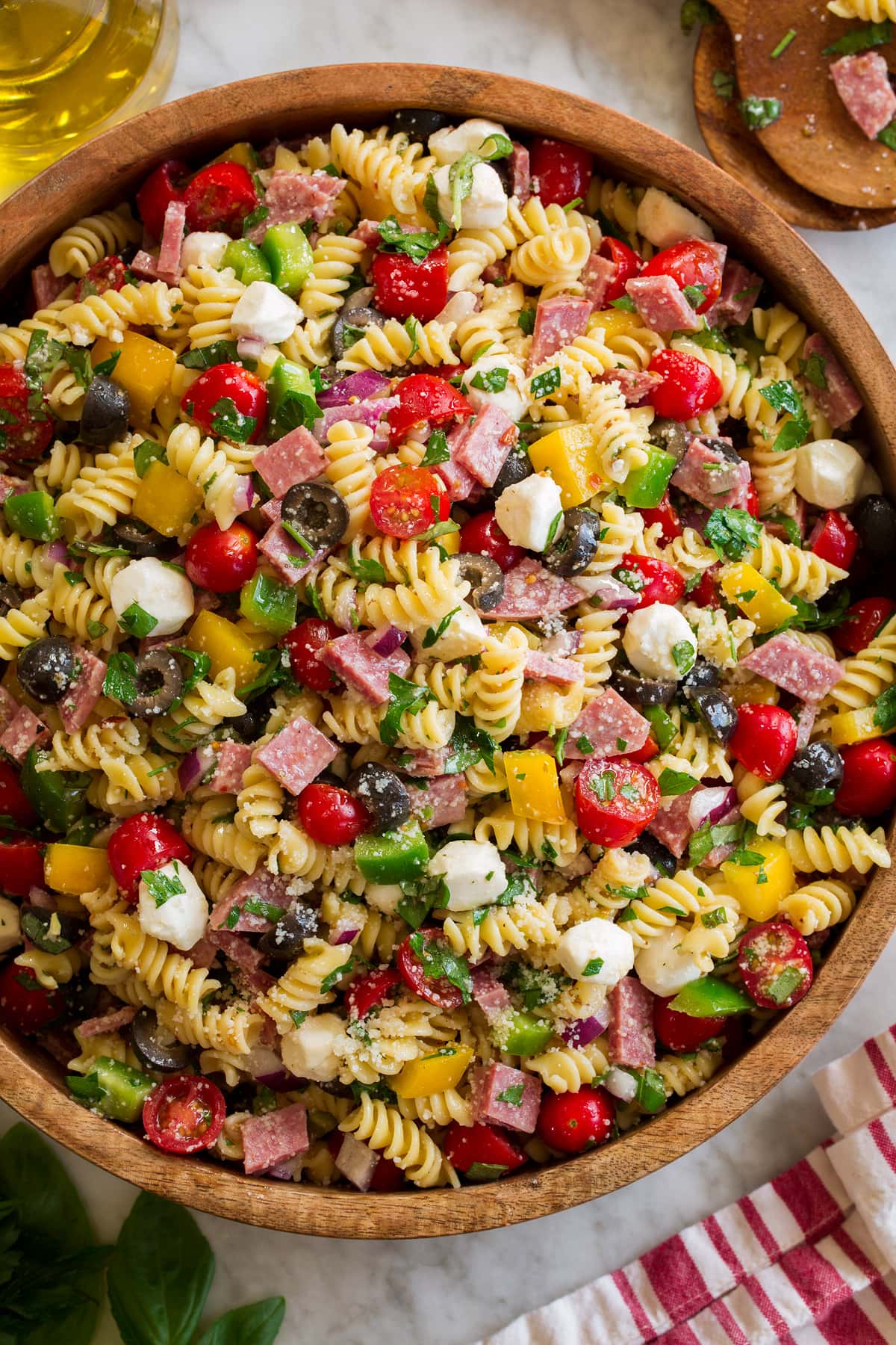 Photo: Italian Pasta Salad shown in a large wooden bowl from above. Bowl is resting on a marble surface with a striped cloth and jar of olive oil to the side.