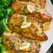 Four pieces of lemon pepper coated chicken aligned on a white oval platter with steamed broccoli to the side.