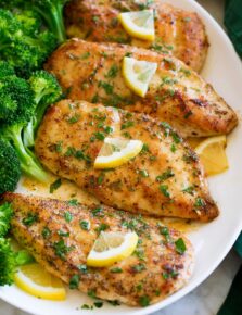 Four pieces of lemon pepper coated chicken aligned on a white oval platter with steamed broccoli to the side.