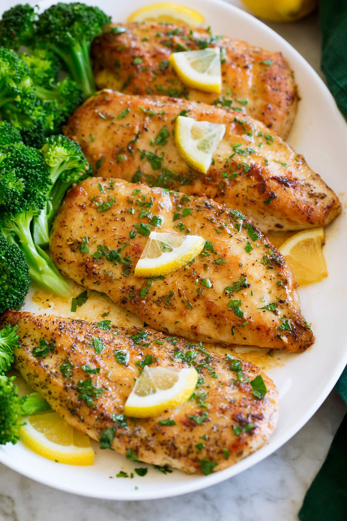 Four pieces of lemon pepper coated chicken aligned on a white oval platter with steamed broccoli to the side.