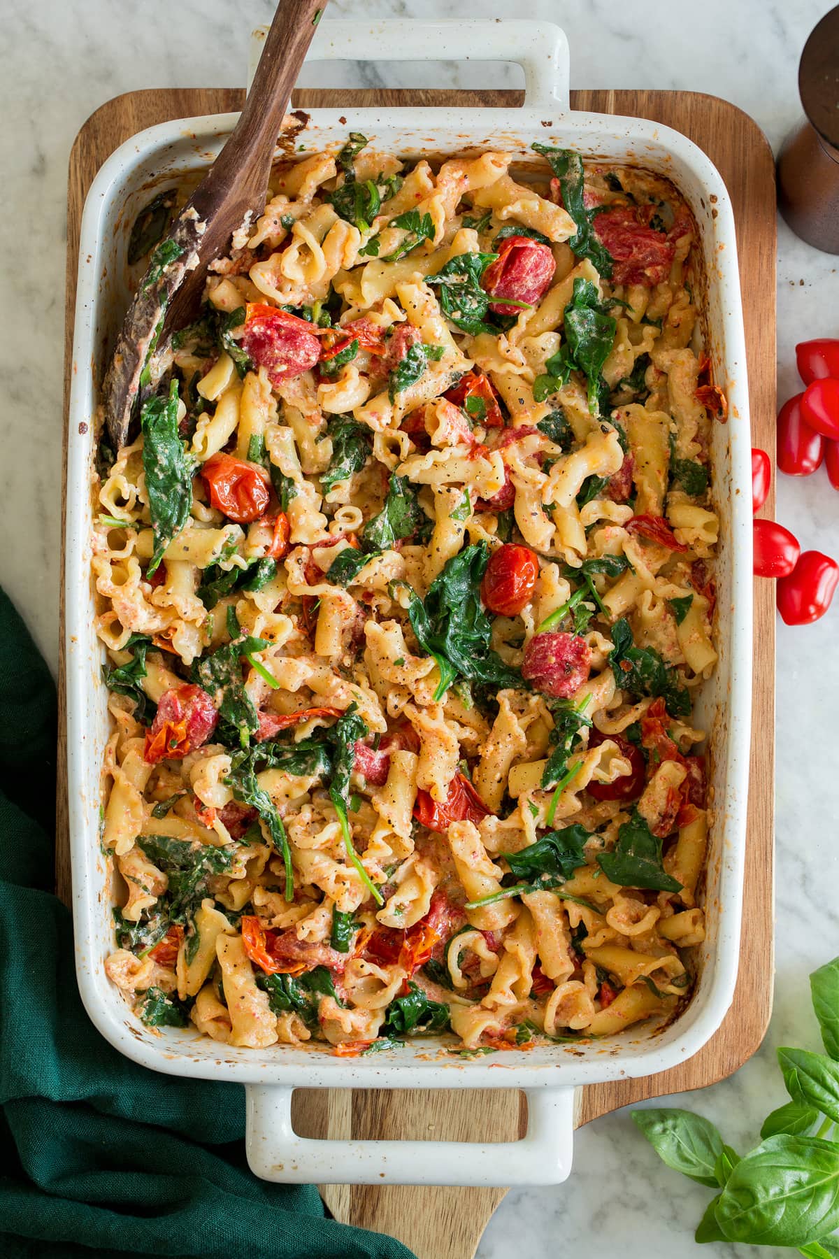 Photo: TikTok Baked Feta Pasta shown in a white baking dish on a wooden board over a white marble surface with a green cloth to the side.