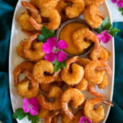 Photo: Many coconut shrimp spread out on a white oval platter with mango sauce in the center in a wooden cup. Vibrant pink flowers are dotted throughout for decoration and platter is resting on a blue cloth over a marble surface.