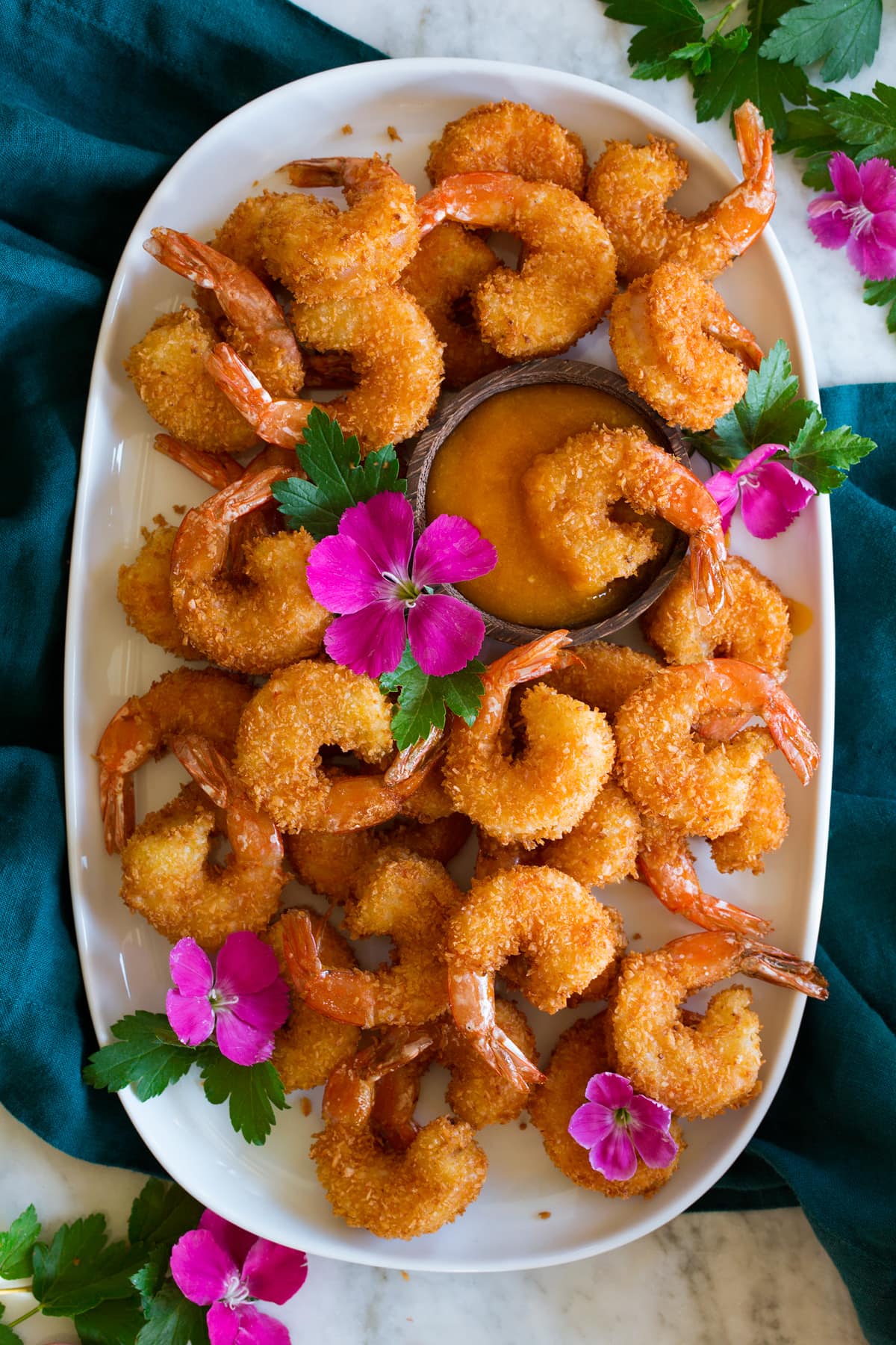 Photo: Many coconut shrimp spread out on a white oval platter with mango sauce in the center in a wooden cup. Vibrant pink flowers are dotted throughout for decoration and platter is resting on a blue cloth over a marble surface.