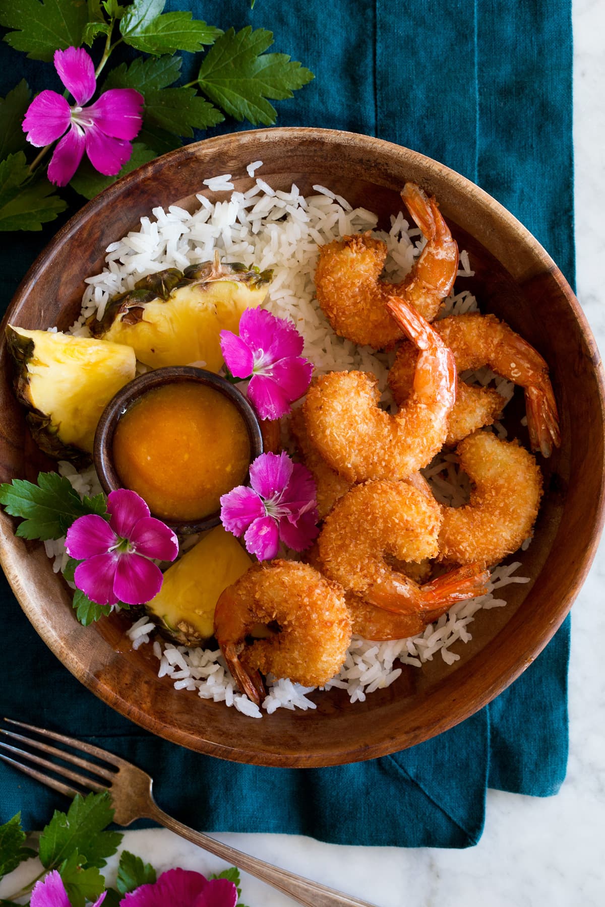 Photo: Coconut shrimp in a wooden bowl over coconut rice. It is served with a side of pineapple and bright pink flowers to the side.