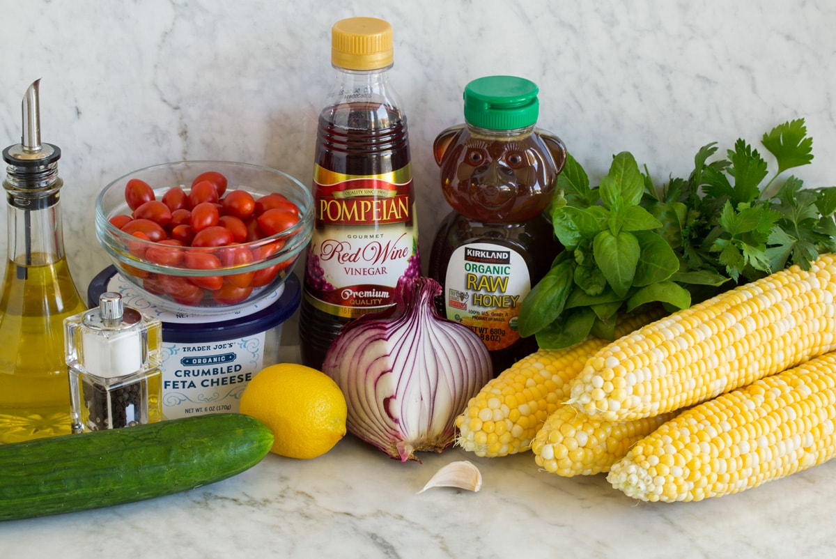 Photo: Ingredients used to make corn salad shown in image. Includes corn on the cob, cucumber, red onion, parsley, basil, tomatoes, feta, red wine vinegar, lemon, honey, olive oil, garlic, salt and pepper.