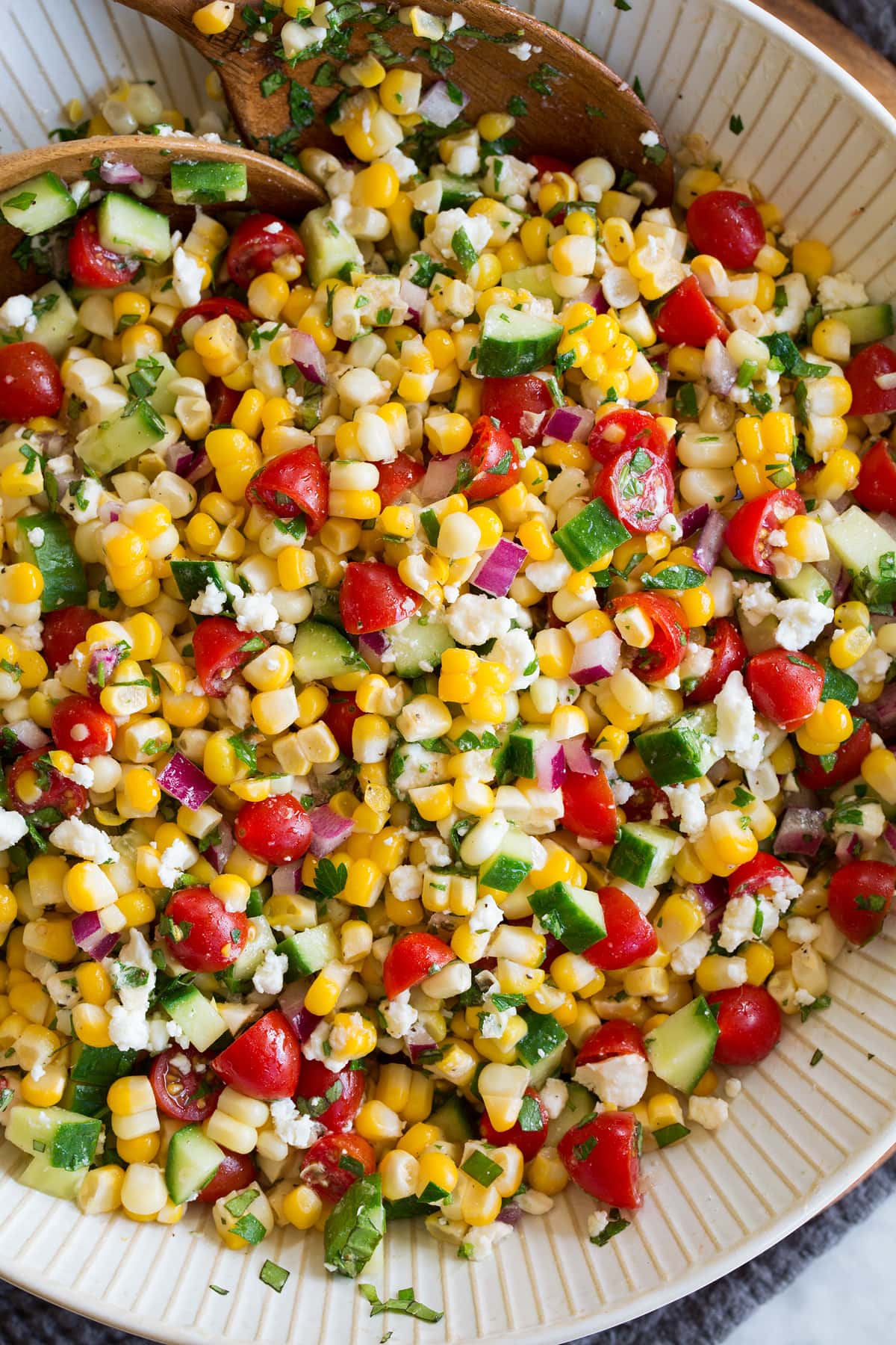 Photo: Corn salad shown close up from above in a serving bowl with wooden serving spoons.