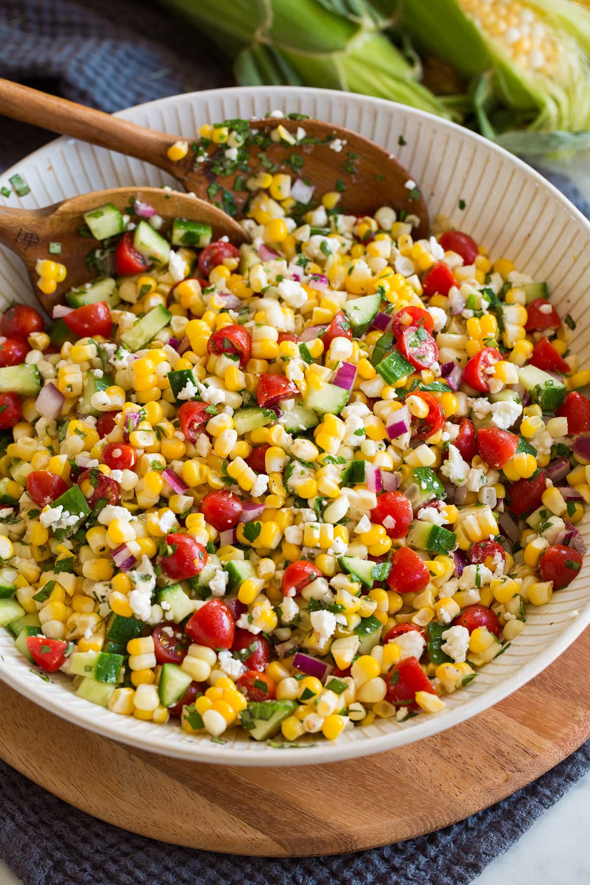 Photo: Corn salad shown in a white serving bowl over a wooden platter and blue cloth. Salad includes fresh corn, tomatoes, red onion, cucumber, feta, herbs and dressing.