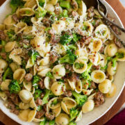 Photo: Orecchiette pasta with sausage and broccoli in a white serving bowl set over a wooden platter on a marble surface. It is shown from above.