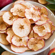 Apple chips shown in a white bowl on a wooden tabletop with fresh apples and leaves surrounding them.