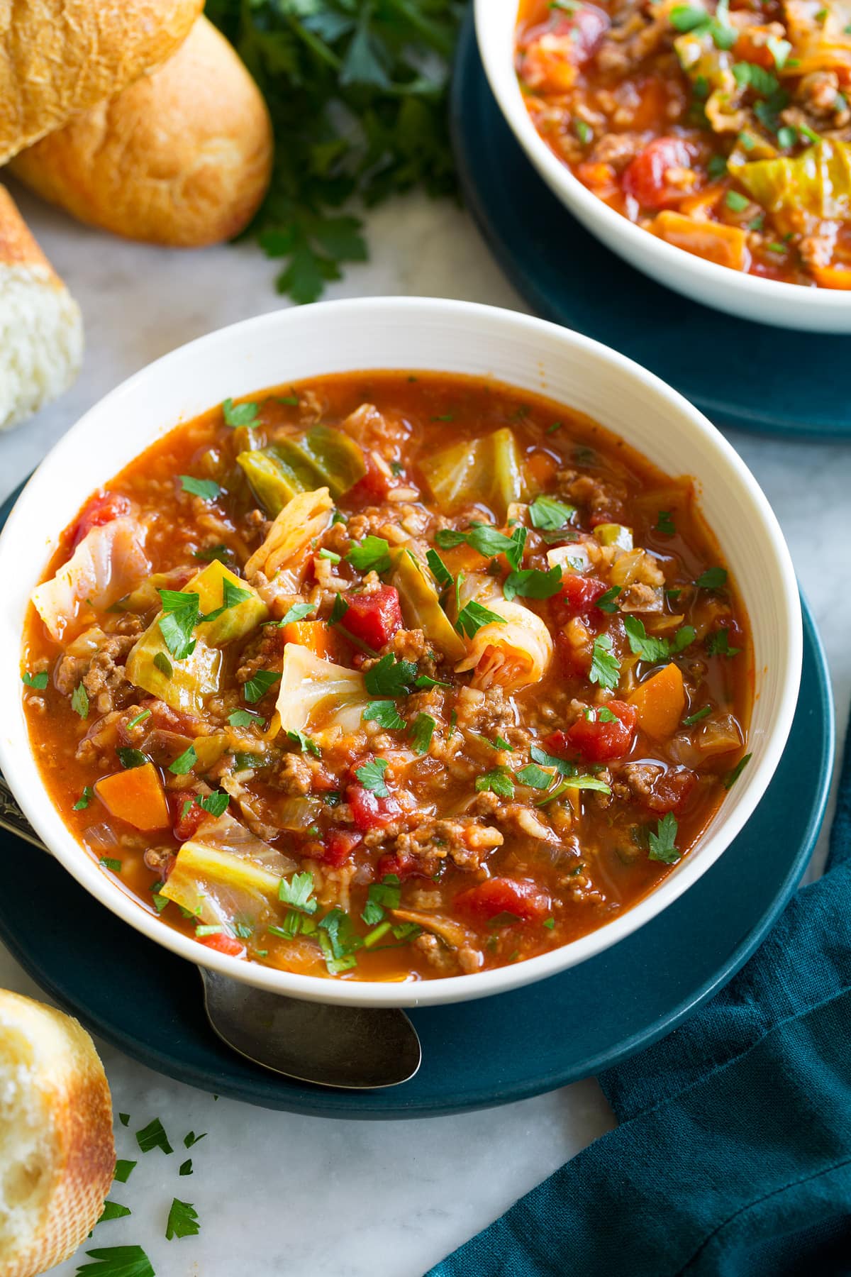 Single serving of cabbage roll soup in a white bowl set over a blue plate with fresh bread shown as a serving suggestion on the side.