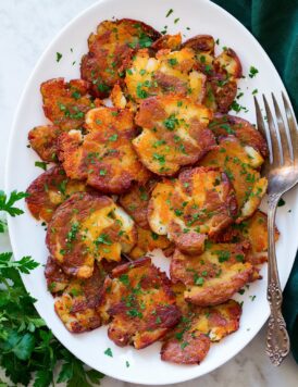 Smashed red potatoes shown piled on a white oval platter. They are shown from above on a white marble surface with a green cloth and parsley to the sides.