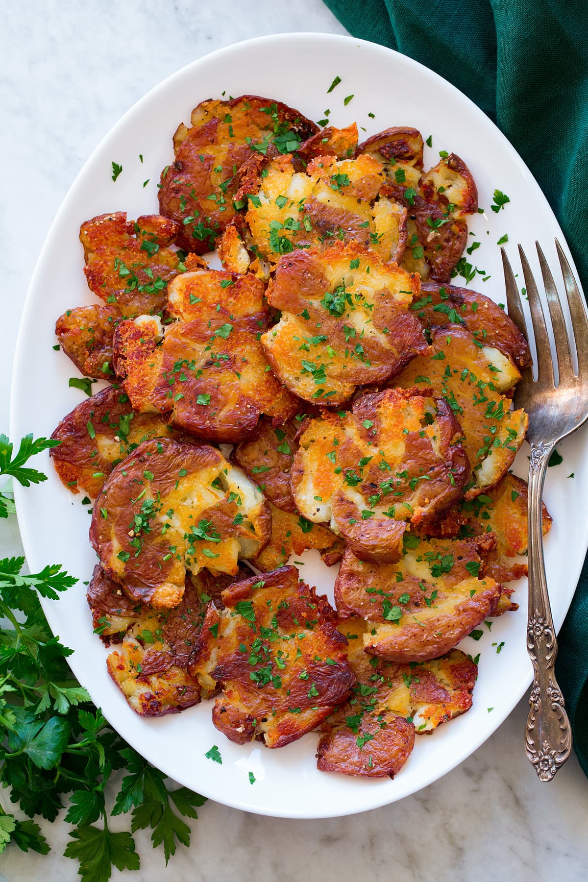 Smashed red potatoes shown piled on a white oval platter. They are shown from above on a white marble surface with a green cloth and parsley to the sides.