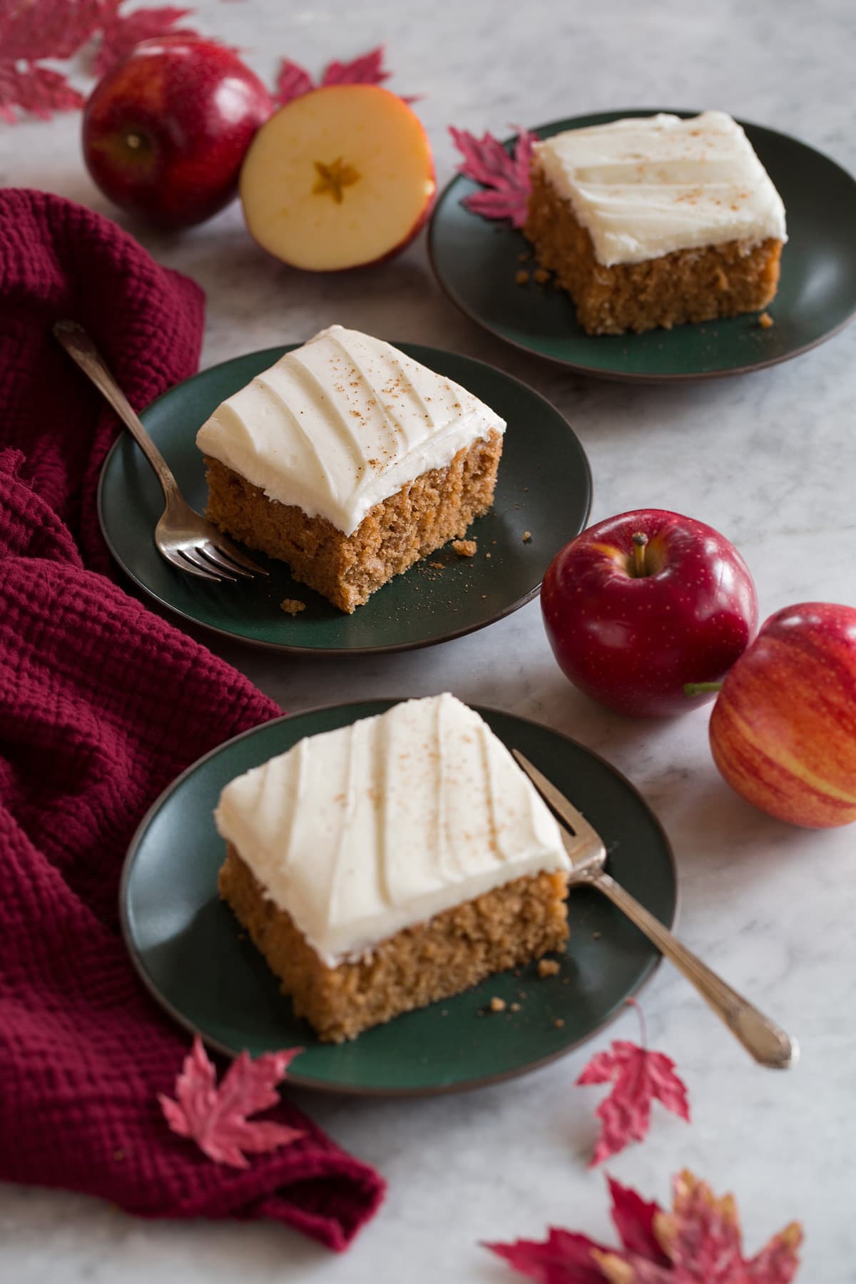 Three slices of applesauce cake shown on green dessert plates with apples, autumn leaves, and a red cloth to the sides.