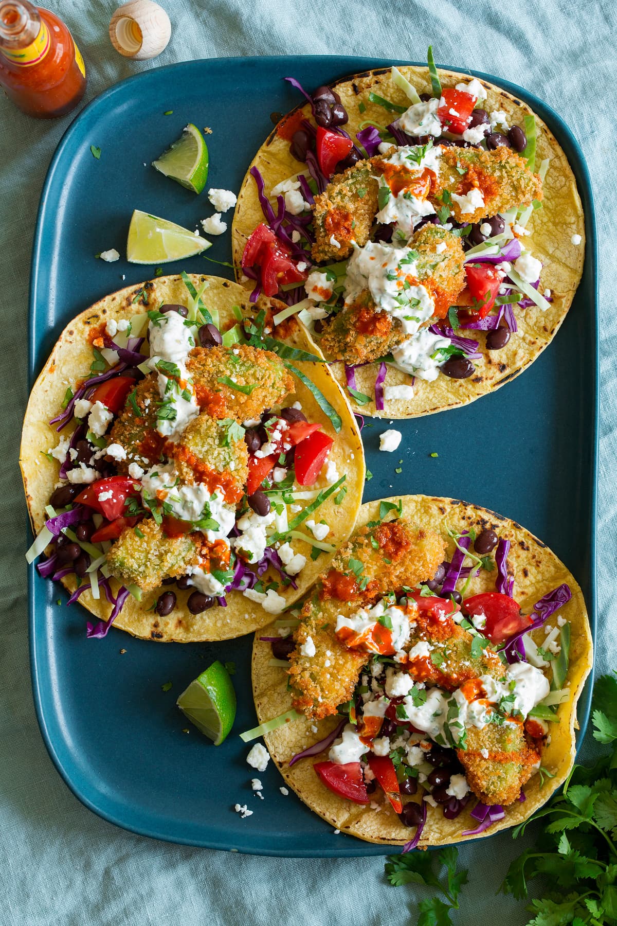 Three avocado tacos shown on a blue platter from above. Tacos are made with avocado fries, tomatoes, cabbage, cilantro sauce, cheese and black beans.