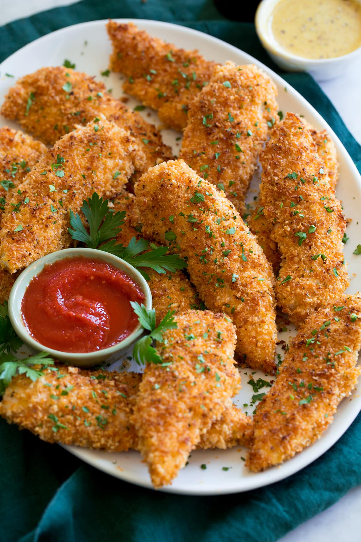 Baked chicken tenders shown on a white oval serving platter over a blue cloth on a marble surface with dipping sauces.