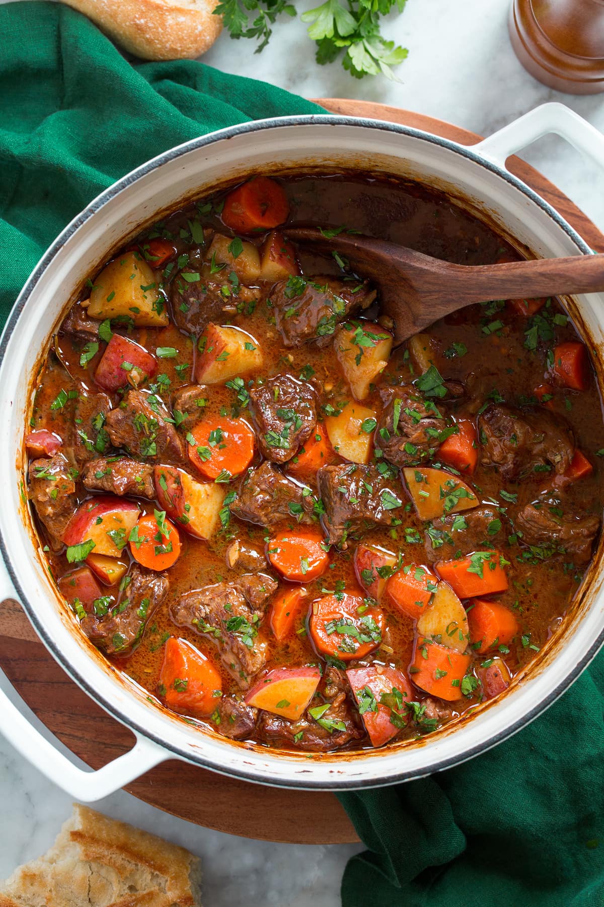 Beef stew shown in a large white pot from overhead. It is resting on a green cloth over a wooden platter.