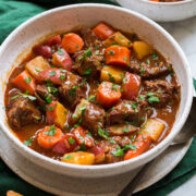 Beef Stew shown in a white serving bowl over a green cloth. Bread is shown to the side as a serving suggestion.