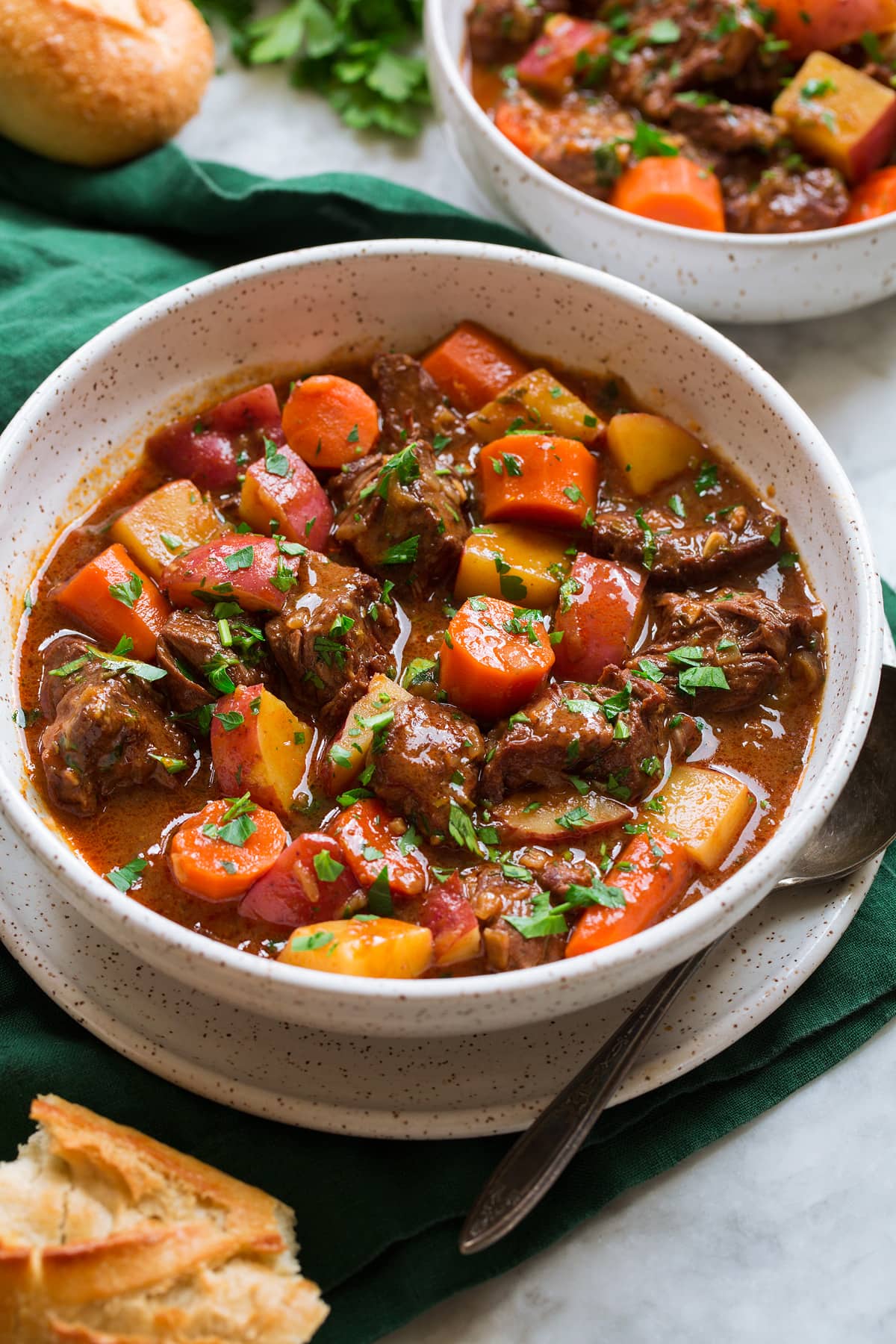 Beef Stew shown in a white serving bowl over a green cloth. Bread is shown to the side as a serving suggestion.