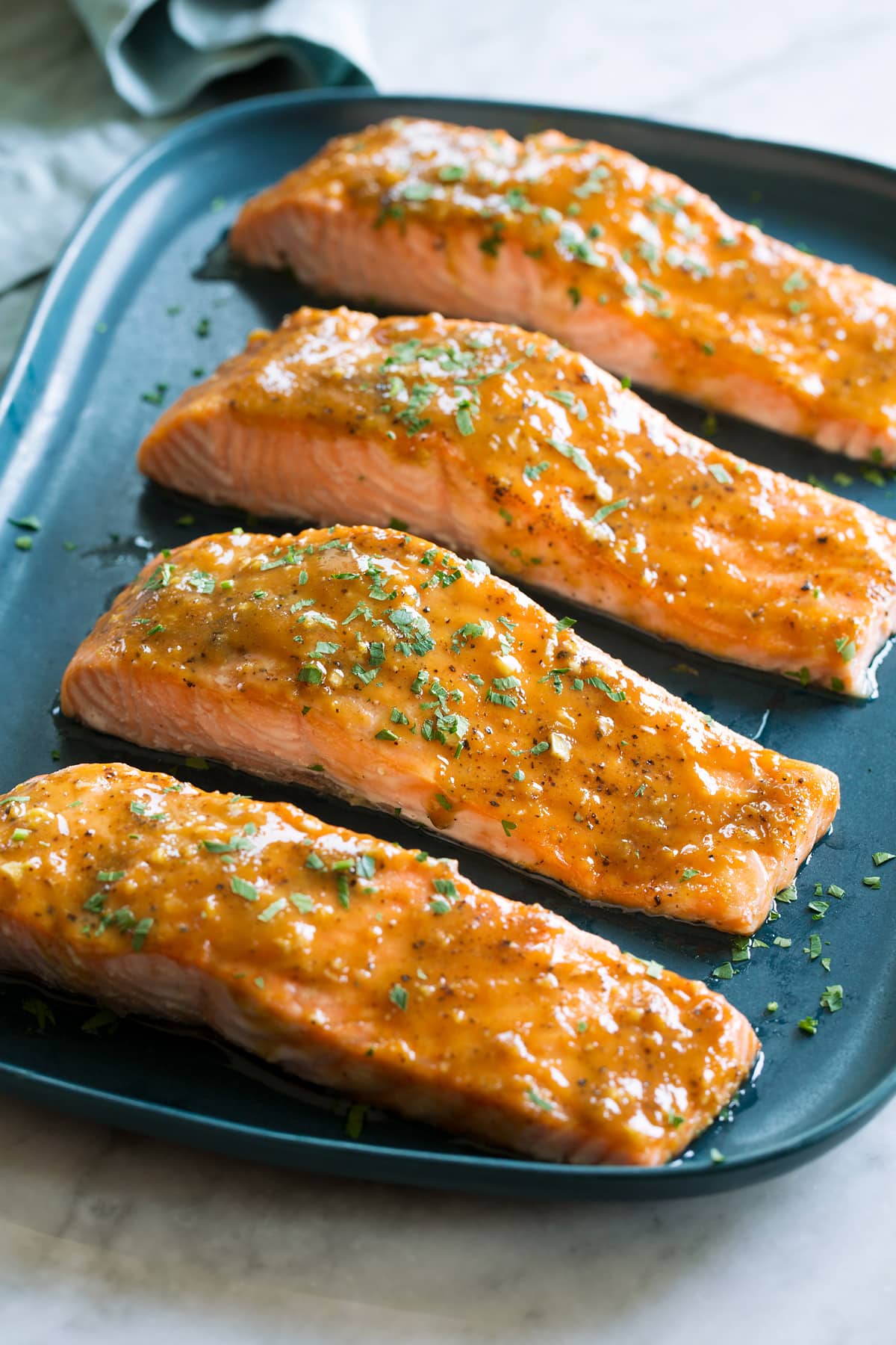 Photo of four salmon fillets coated with a brown sugar glaze resting on a blue rectangular platter on a white marble surface.