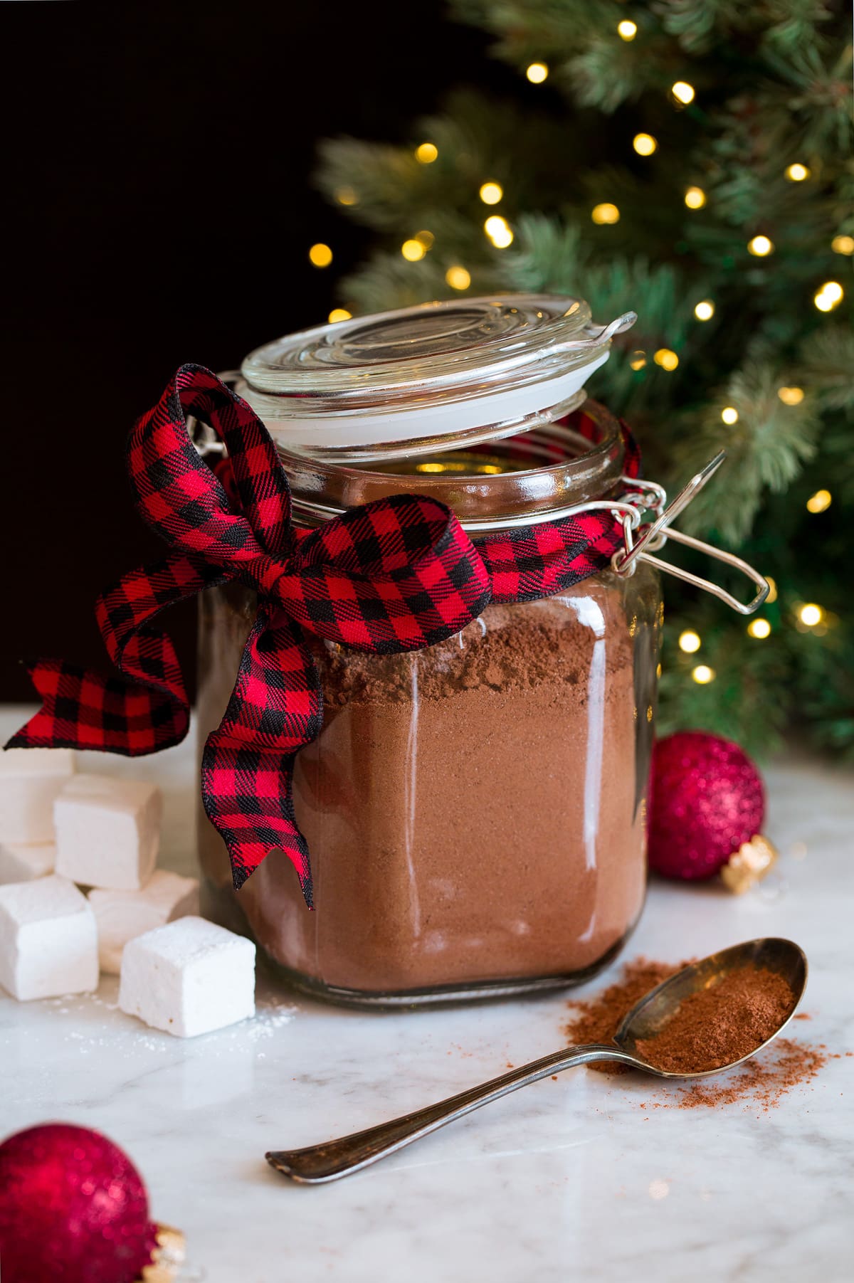 Homemade hot chocolate mix in a glass canister with a buffalo check red and black ribbon. A Christmas tree is in the background.