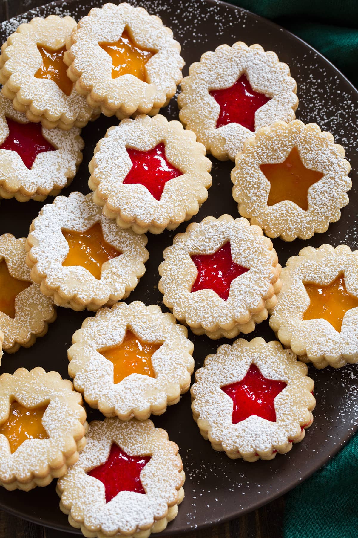 Powdered sugar dusted linzer cookies filled with fruit jam shown on a dark brown plate.