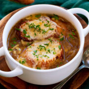 French onion soup shown in a white bowl with handles. Soup is topped with baguette slices, broiled cheese and herbs.