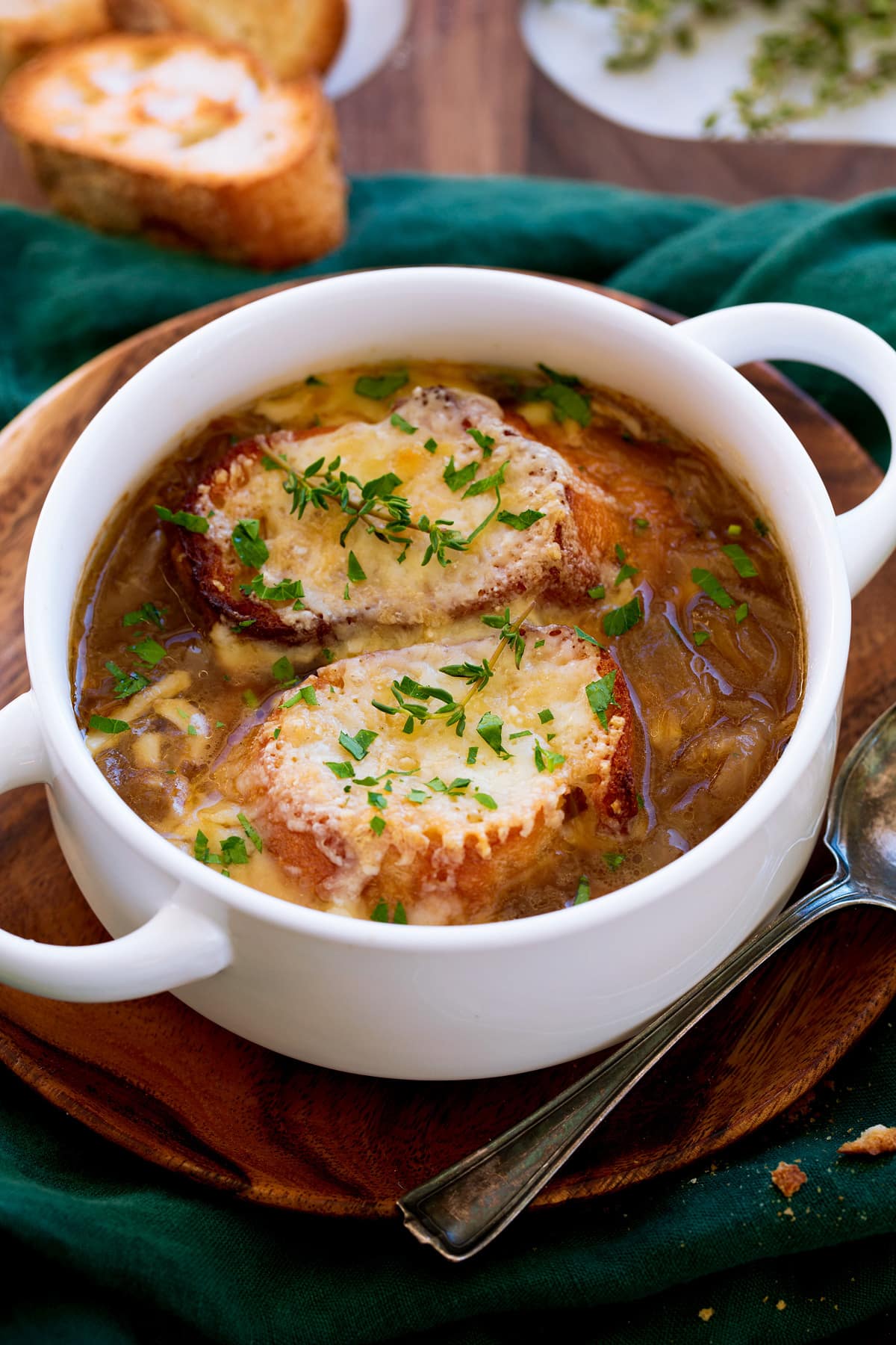 French onion soup shown in a white bowl with handles. Soup is topped with baguette slices, broiled cheese and herbs.