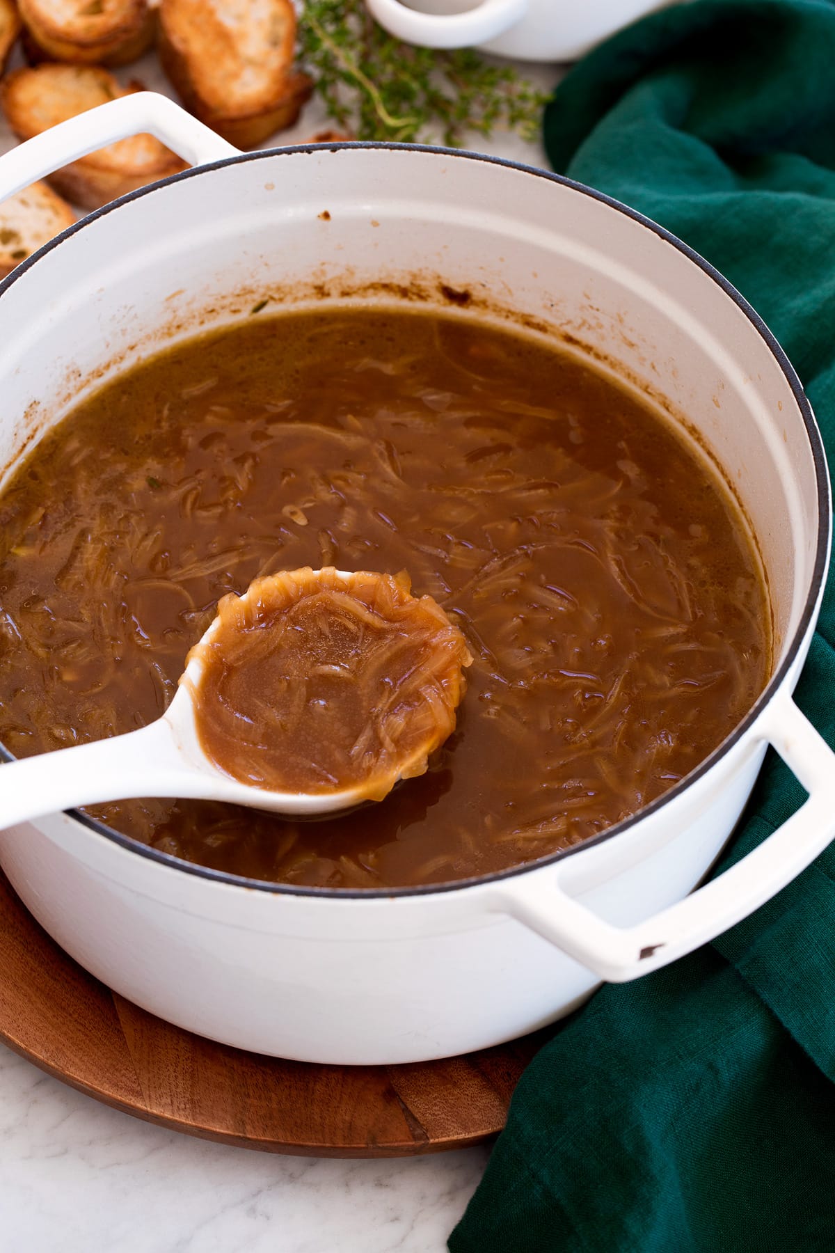 Large white pot filled with french onion soup. A ladle is scooping soup and pot is resting on a wooden platter with a green cloth.