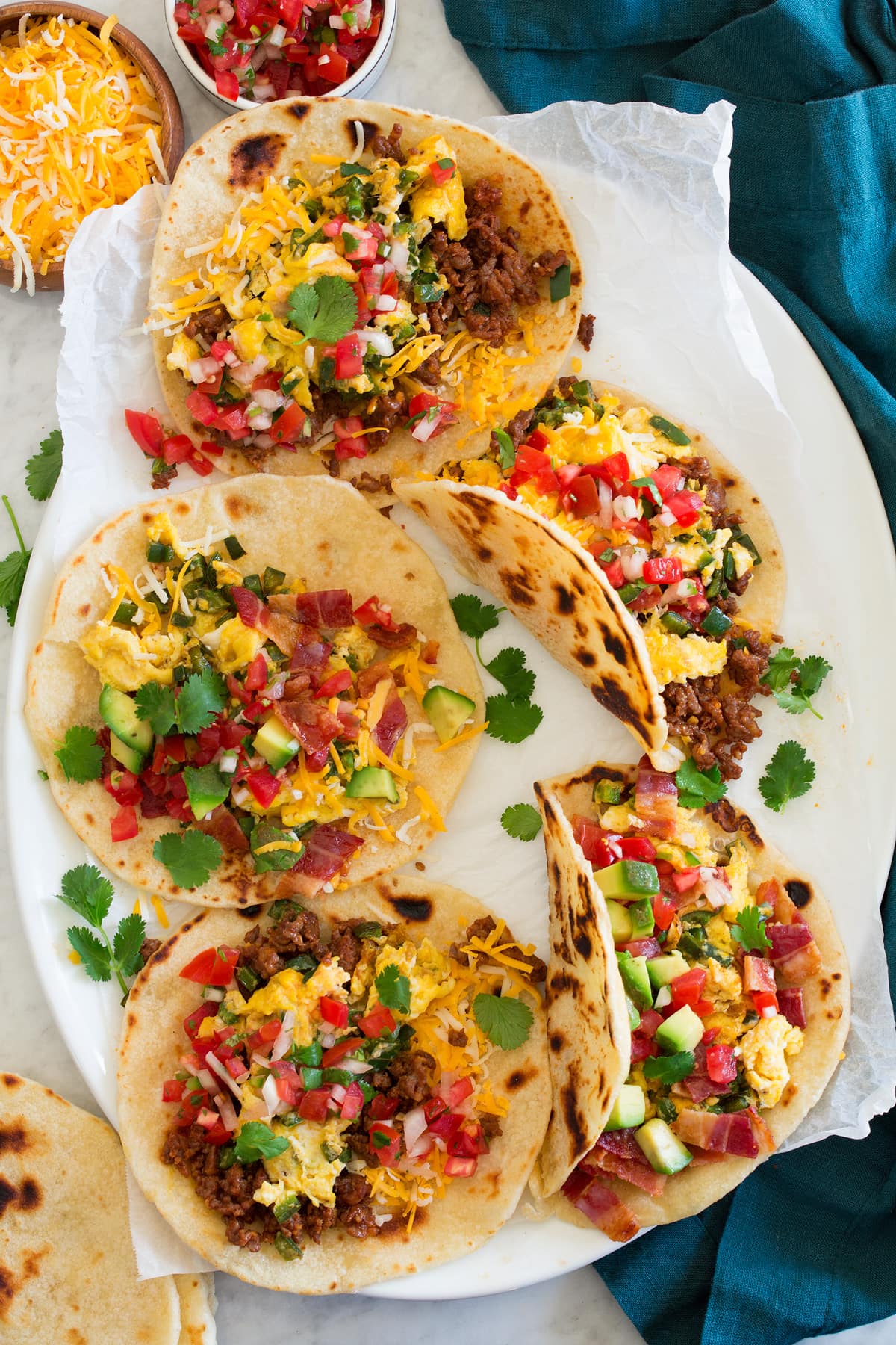 Breakfast tacos made with flour tortillas, chorizo, bacon, eggs, cheese, avocado and pico de gallo shown on a white oval platter from above.