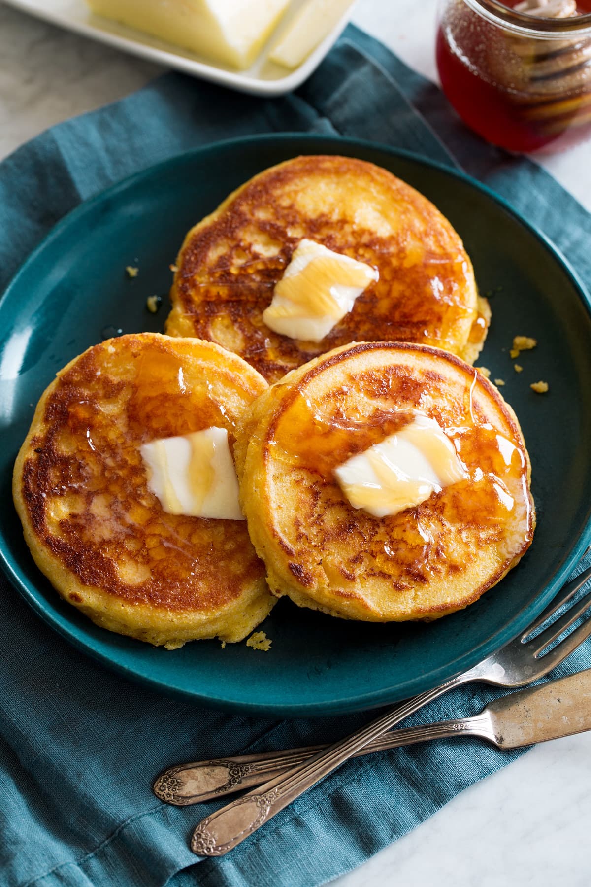 Three johnny cakes covered with dabs of butter and honey. They are a blue plate set over a blue cloth with silverware to the side.