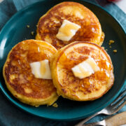 Three johnny cakes covered with dabs of butter and honey. They are a blue plate set over a blue cloth with silverware to the side.