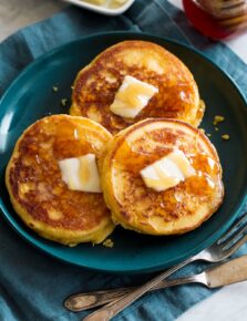 Three johnny cakes covered with dabs of butter and honey. They are a blue plate set over a blue cloth with silverware to the side.