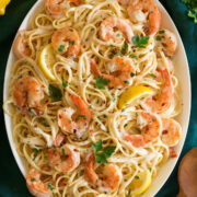 Shrimp pasta with creamy lemon sauce on a white oval serving platter shown from above. Platter is resting on an aqua colored cloth and lemons are shown to the side.