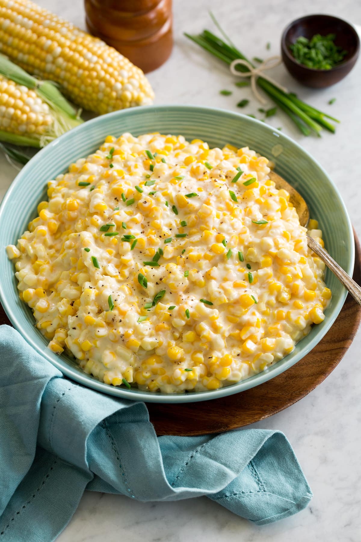 Creamed corn in a turquoise serving bowl garnished with chives and pepper. Bowl is resting on a wood plate with a blue cloth to the side.