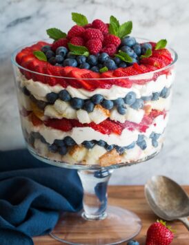 Layered trifle in a glass trifle dish. It includes cubes of angel food cake, blueberries, strawberries, raspberries and a cream cheese whipped cream topping.